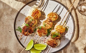 Prepare a grill for medium-high heat. Mix mayonnaise, lime juice, a pinch of salt, and 1 Tbsp. water in a small bowl; set lime mayo aside.
Finely grind nori in spice mill. Transfer half to a small bowl; set aside for serving. Transfer remaining nori to a large bowl and mix in coriander, ginger, and 2 Tbsp. oil. Add scallops and toss to coat.
Thread 3 scallops onto 2 skewers. (This will keep scallops in place and make them easy to turn. You can also use this method for shrimp and small peppers like shishito and Padrón.) Repeat with remaining scallops and skewers. Season both sides with salt.
Clean and oil grate, then immediately place scallops on the outside edge of the grill so that the skewers are hanging off the side. Grill, turning scallops with handles of skewers, until grill marks appear and scallops are just cooked through, about 3 minutes per side, depending on their size.
Spread lime mayo on a platter and place skewers with scallops on top. Finely grate zest from lime half over, then squeeze juice over. Top with scallions and sprinkle with Aleppo-style pepper and reserved nori.